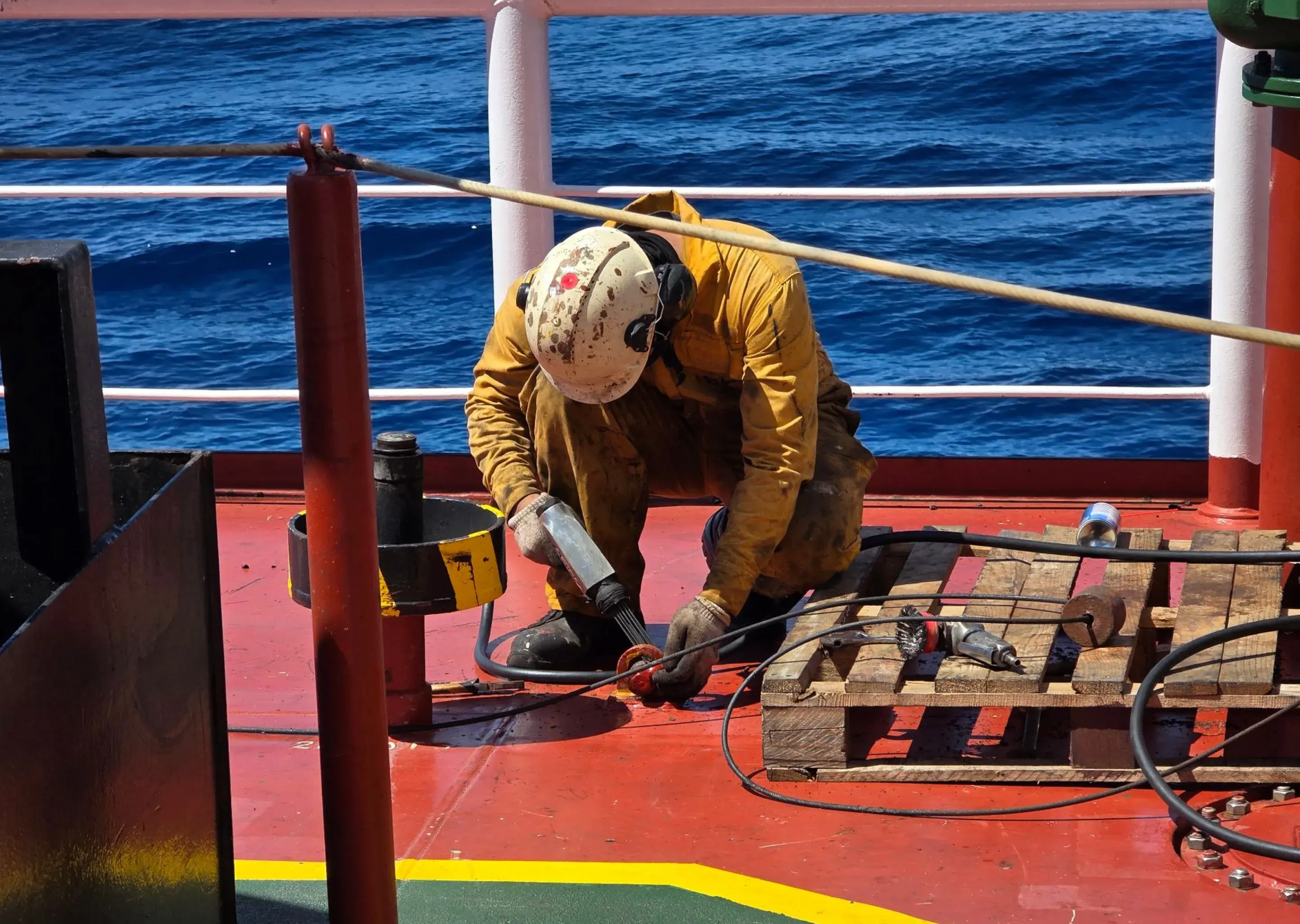 Crew member in a hard hat and coveralls carrying out maintenance on a vessel deck at sea